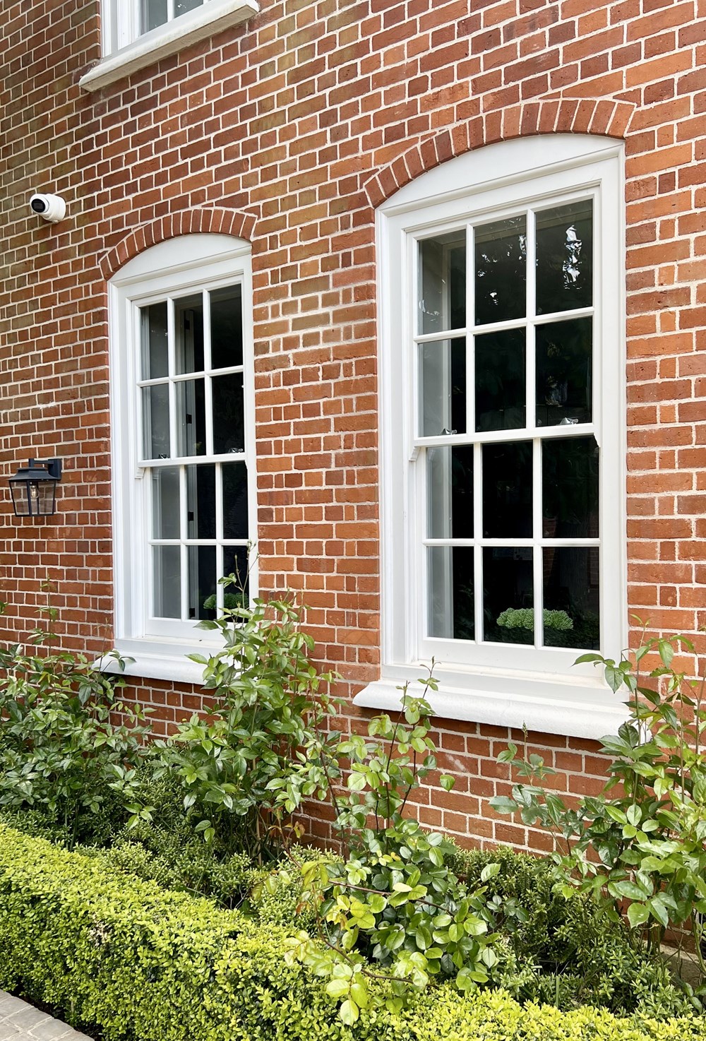 Exterior shot of white painted Timber sliding sash windows installed on a brick building with green hedges and shrubbery in front of the windows. 